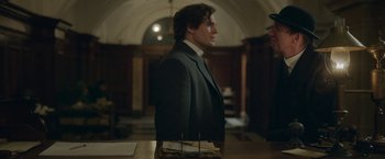 Movie still from “Enola Holmes 2” (2022), directed by Harry Bradbeer – A man in a suit standing in front of a table; Medium shot, Over the shoulder angle