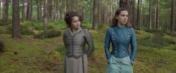 Movie still from “Enola Holmes 2” (2022), directed by Harry Bradbeer – Two women standing next to each other in a wooded area; Medium shot, Over the shoulder angle