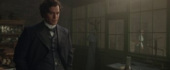 Movie still from “Enola Holmes 2” (2022), directed by Harry Bradbeer – A man in a suit and tie standing next to a window; Medium shot, Over the shoulder angle
