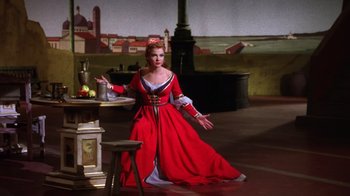 Movie still from “Kiss Me Kate” (1953), directed by George Sidney – A woman dressed in a red dress standing in front of a table; Wide shot, High angle