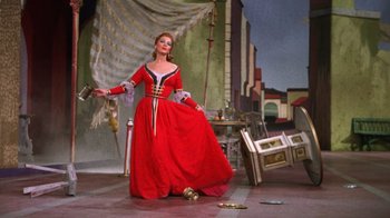 Movie still from “Kiss Me Kate” (1953), directed by George Sidney – A woman in a red dress standing in front of a table; Wide shot, High angle