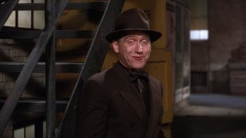 Movie still from “Kiss Me Kate” (1953), directed by George Sidney – A man in a suit and hat standing in front of a staircase; Medium shot, Low angle
