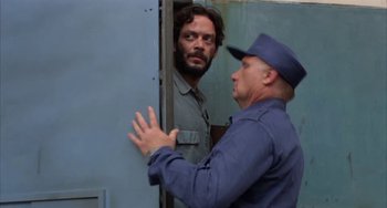 Movie still from “Kiss of the Spider Woman” (1985), directed by Hector Babenco – A man standing next to another man near a door; Medium shot, Over the shoulder angle