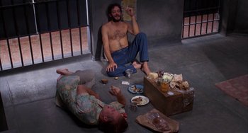 Movie still from “Kiss of the Spider Woman” (1985), directed by Hector Babenco – A man sitting on the ground next to another man; Wide shot, High angle