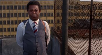 Movie still from “Kiss of the Spider Woman” (1985), directed by Hector Babenco – A man in a vest and tie standing in front of a fence; Medium shot, Low angle