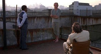 Movie still from “Kiss of the Spider Woman” (1985), directed by Hector Babenco – A man standing on a balcony next to two other people; Wide shot, Over the shoulder angle