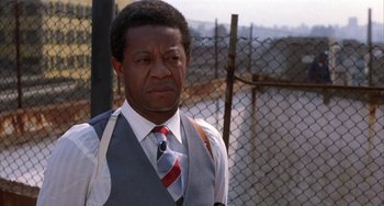 Movie still from “Kiss of the Spider Woman” (1985), directed by Hector Babenco – A man wearing a vest and tie standing in front of a chain link fence; Close Up shot, Low angle