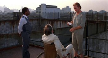 Movie still from “Kiss of the Spider Woman” (1985), directed by Hector Babenco – A man standing next to two other men in a wheelchair; Wide shot, Over the shoulder angle