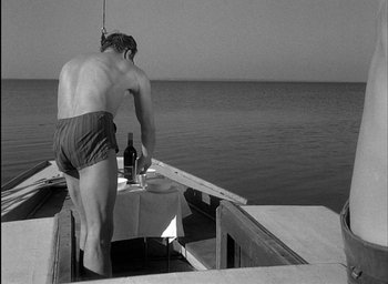 Movie still from “Knife in the Water” (1962), directed by Roman Polanski – A man standing on the side of a boat with a bottle of wine; Wide shot, Low angle
