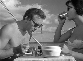 Movie still from “Knife in the Water” (1962), directed by Roman Polanski – A man and a woman are sitting at a table with food; Medium shot, Low angle