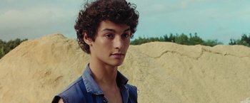Movie still from “Knife + Heart” (2018), directed by Yann Gonzalez – A man with curly hair is standing in front of some rocks; Close Up shot, Low angle