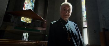 Movie still from “Knight of Cups” (2015), directed by Terrence Malick – An older man wearing a priest's outfit and holding a cross; Medium shot, Low angle