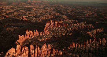 Movie still from “Koyaanisqatsi” (1982), directed by Godfrey Reggio – An aerial view of a desert landscape with rocks and trees; Extreme Wide shot, High angle