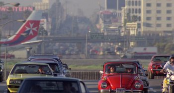 Movie still from “Koyaanisqatsi” (1982), directed by Godfrey Reggio – A red vw bug driving down a street next to a busy highway; Extreme Wide shot, High angle