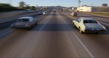 Movie still from “Koyaanisqatsi” (1982), directed by Godfrey Reggio – Cars driving down a highway with a mountain in the background; Wide shot, High angle