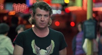 Movie still from “Koyaanisqatsi” (1982), directed by Godfrey Reggio – A man with long hair wearing a t - shirt with an eagle on it; Close Up shot, Low angle