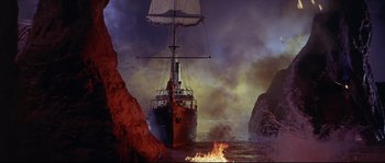 Movie still from “Krakatoa: East of Java” (1968), directed by Bernard L. Kowalski – A boat in the water near some rocks and a fire; Extreme Wide shot, High angle