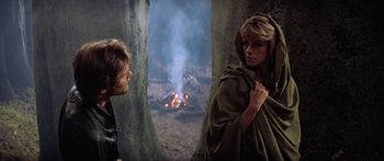 Movie still from “Krull” (1983), directed by Peter Yates – A woman in a hooded cloak standing in front of a fire; Medium shot, Over the shoulder angle