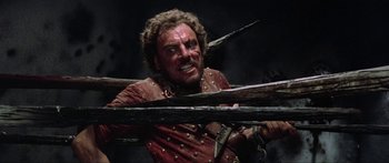Movie still from “Krull” (1983), directed by Peter Yates – A man in a red shirt is holding a wooden stick; Close Up shot, Low angle