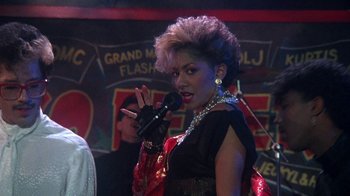 Movie still from “Krush Groove” (1985), directed by Michael Schultz – A woman in a black and red dress holding a microphone; Close Up shot, Over the shoulder angle