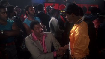 Movie still from “Krush Groove” (1985), directed by Michael Schultz – Two men are shaking hands in a crowded room; Medium shot, Over the shoulder angle