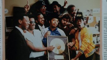 Movie still from “Krush Groove” (1985), directed by Michael Schultz – A group of men holding up a gold record; Medium shot, Overhead angle