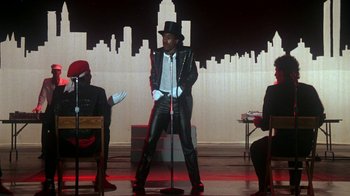 Movie still from “Krush Groove” (1985), directed by Michael Schultz – A man in a black leather jacket and hat on a stage; Medium shot, Over the shoulder angle