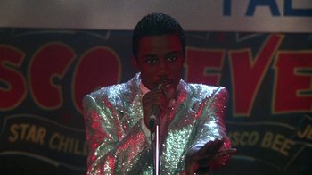 Movie still from “Krush Groove” (1985), directed by Michael Schultz – A man in a silver jacket is holding a microphone; Medium shot, Over the shoulder angle