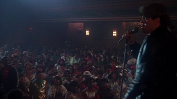 Movie still from “Krush Groove” (1985), directed by Michael Schultz – A crowd of people sitting and standing in front of a microphone; Wide shot, Over the shoulder angle