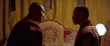 Movie still from “Kundun” (1997), directed by Martin Scorsese – Two men are standing next to each other in front of a chair; Medium shot, Over the shoulder angle