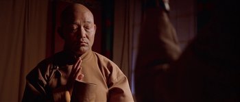 Movie still from “Kundun” (1997), directed by Martin Scorsese – An older bald man in a tan shirt is holding his hands to his chest; Close Up shot, Low angle