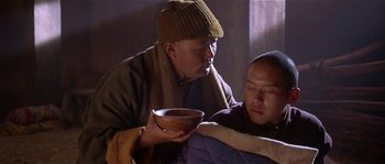 Movie still from “Kundun” (1997), directed by Martin Scorsese – A man holding a wooden bowl next to another man; Medium shot, High angle