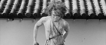 Movie still from “Black Cat” (1968), directed by Kaneto Shindô – A shirtless man with long dreadlocks is walking outside; Medium shot, Low angle