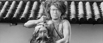 Movie still from “Black Cat” (1968), directed by Kaneto Shindô – A man with long hair is holding a knife in his mouth and a man with long hair is holding a knife in his mouth; Medium shot, Low angle