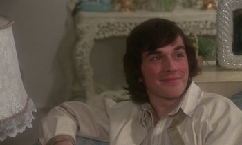 Movie still from “La Cage aux Folles” (1978), directed by Édouard Molinaro – A man sitting in a chair smiling at the camera; Close Up shot, Over the shoulder angle