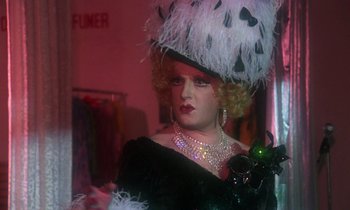 Movie still from “La Cage aux Folles” (1978), directed by Édouard Molinaro – A woman in a black dress wearing a black hat; Close Up shot, Over the shoulder angle
