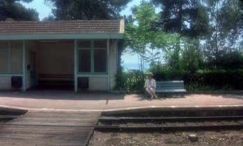 Movie still from “La Cage aux Folles” (1978), directed by Édouard Molinaro – A person sitting on a bench near a building; Extreme Wide shot, High angle