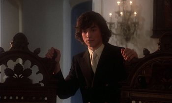 Movie still from “La Cage aux Folles” (1978), directed by Édouard Molinaro – A man in a suit and tie standing next to a chair; Medium shot, Low angle