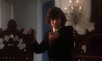 Movie still from “La Cage aux Folles” (1978), directed by Édouard Molinaro – A man in a suit and tie standing next to a wooden chair; Medium shot, Low angle