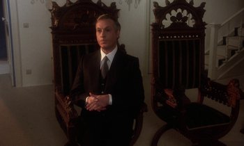 Movie still from “La Cage aux Folles” (1978), directed by Édouard Molinaro – A man wearing a suit and tie sitting in a chair; Medium shot, Low angle