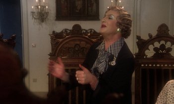 Movie still from “La Cage aux Folles” (1978), directed by Édouard Molinaro – An older woman in a black jacket is singing; Medium shot, High angle