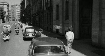 Movie still from “La Notte” (1961), directed by Michelangelo Antonioni – A woman walking down a street past a parked car; Wide shot, High angle