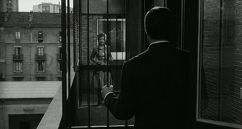 Movie still from “La Notte” (1961), directed by Michelangelo Antonioni – A man standing in front of a woman behind bars; Wide shot, Over the shoulder angle