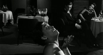 Movie still from “La Notte” (1961), directed by Michelangelo Antonioni – A black and white photo of a woman holding her hands up to her face and a wine glass on top of her head; Medium shot, Over the shoulder angle