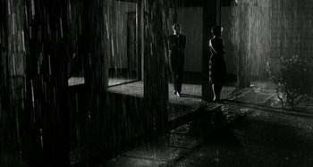 Movie still from “La Notte” (1961), directed by Michelangelo Antonioni – Two people are standing in the rain near a building; Extreme Wide shot, High angle