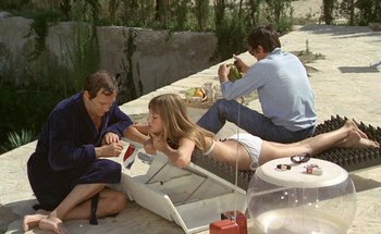 Movie still from “The Swimming Pool” (1969), directed by Jacques Deray – A woman sitting on the ground with two other people; Medium shot, High angle