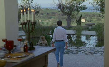Movie still from “The Swimming Pool” (1969), directed by Jacques Deray – A man in a white dress shirt and blue pants walking in front of a table with candles; Wide shot, High angle
