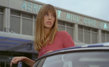Movie still from “The Swimming Pool” (1969), directed by Jacques Deray – A woman standing next to a car in front of an airport; Medium shot, Low angle
