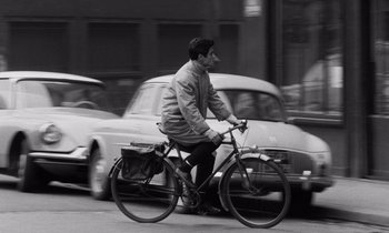 Movie still from “The Truth” (1960), directed by Henri-Georges Clouzot – A man riding a bike down a street next to parked cars; Wide shot, Low angle