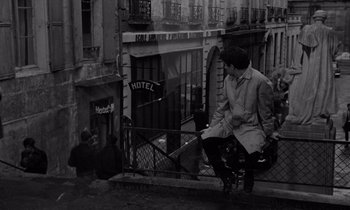 Movie still from “The Truth” (1960), directed by Henri-Georges Clouzot – A man sitting on a fence near a building; Wide shot, Low angle